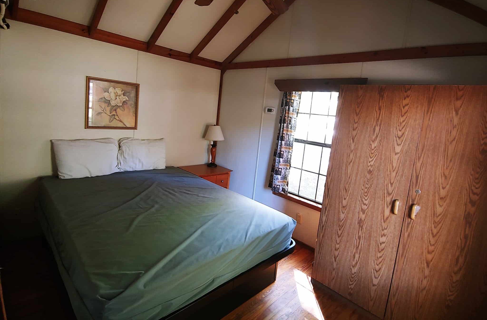 A cozy bedroom featuring a bed with a green bedspread, two pillows, and a wooden headboard. Above the bed is a floral picture. A side table with a lamp sits next to the bed. A large wooden wardrobe stands near a window with patterned curtains. The room has a wooden ceiling fan.