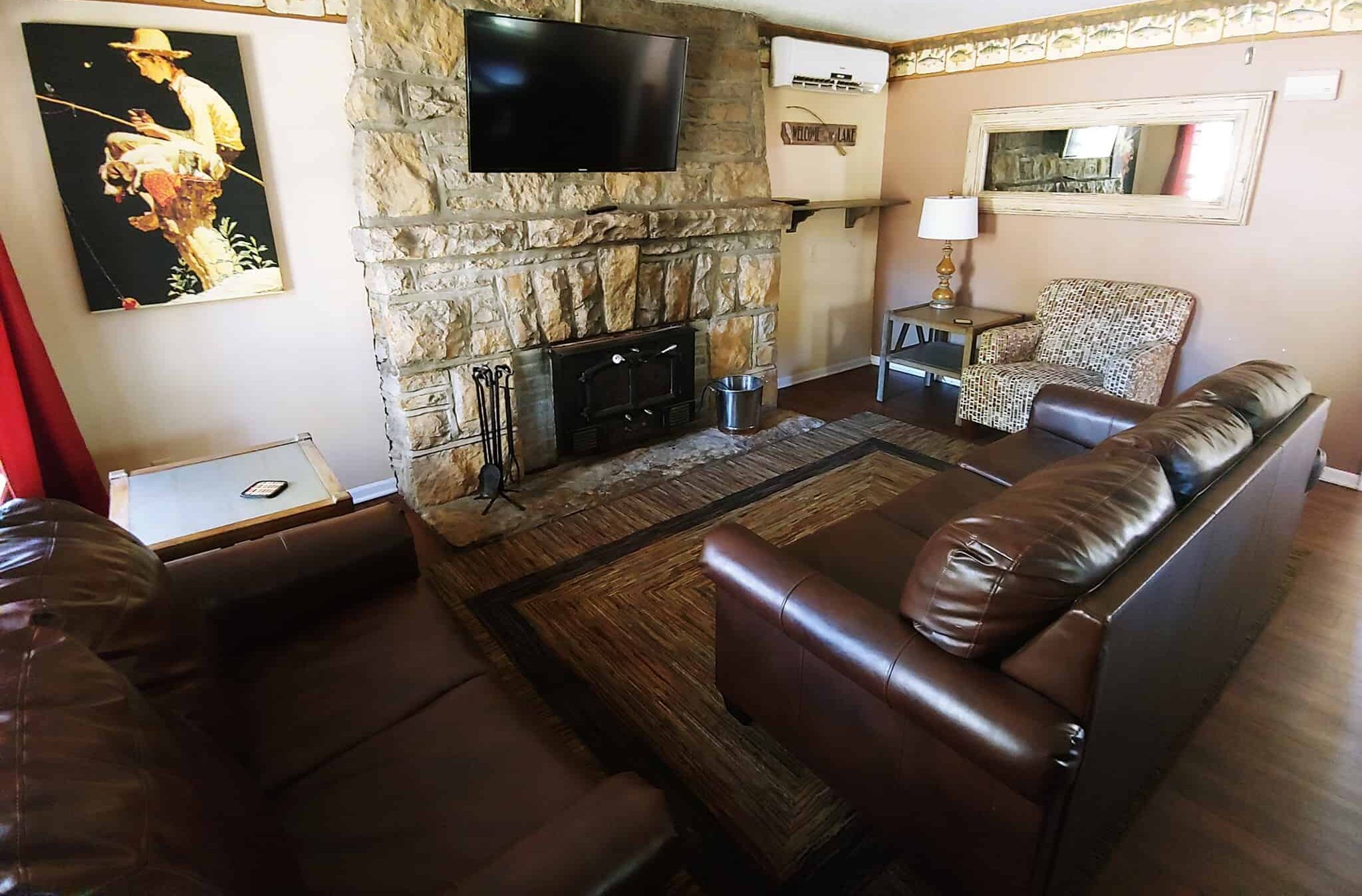 A cozy living room with a stone fireplace, a mounted TV above it, and brown leather couches. A painting of a person fishing hangs on the left wall. A large mirror, a patterned chair, and a table with a lamp are to the right of the fireplace. The room has wooden floors and a fan on the ceiling.