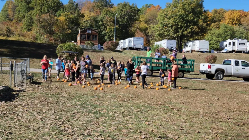 A group of people, including adults and children, gathered in an open field with several small pumpkins scattered on the ground at Basswood Resort. A green tractor with a trailer is parked nearby. Trees with autumn foliage and parked trailers are in the background, capturing the essence of October.