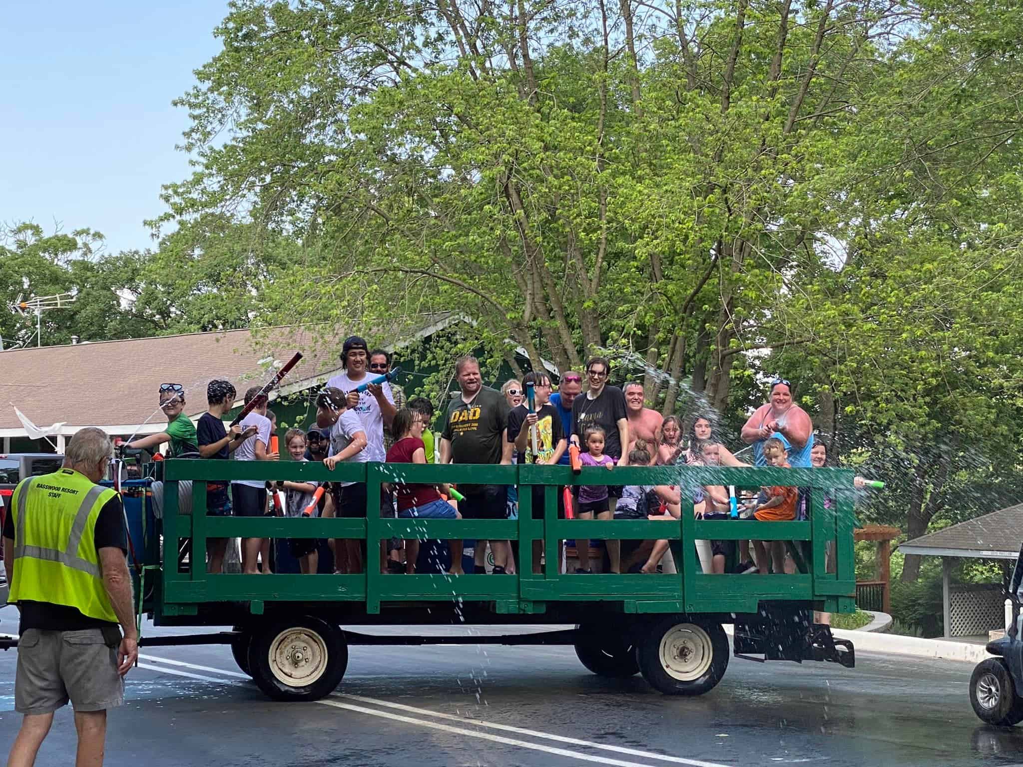 A group of people riding on a green open-sided trailer being towed by a vehicle. Everyone is smiling, waving, and some people are spraying water. They appear to be enjoying a sunny day, with trees in the background and a man in a safety vest walking nearby.
