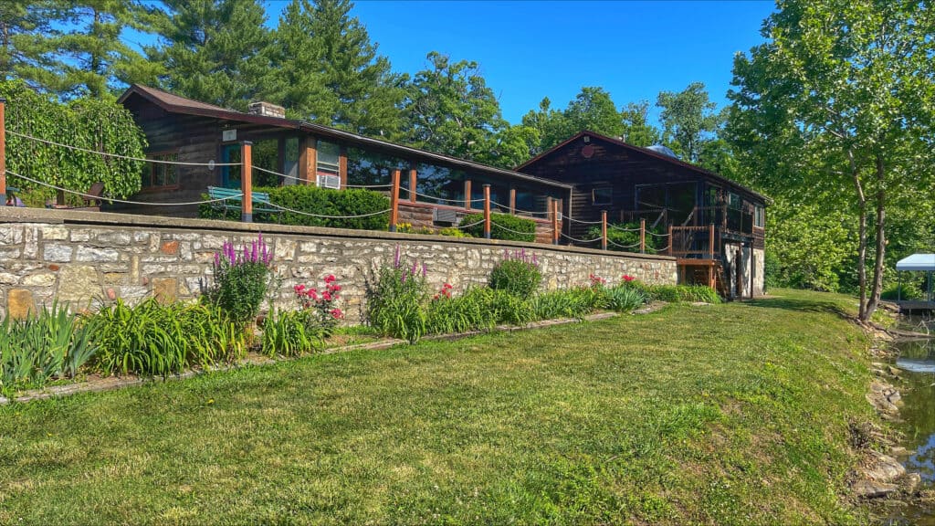 A rustic stone and wood house is surrounded by lush greenery. The home has a long deck with a rope railing, and vibrant flowers bloom below the deck. A grassy lawn leads to a wooded area in the background, and the sky is clear and blue.
