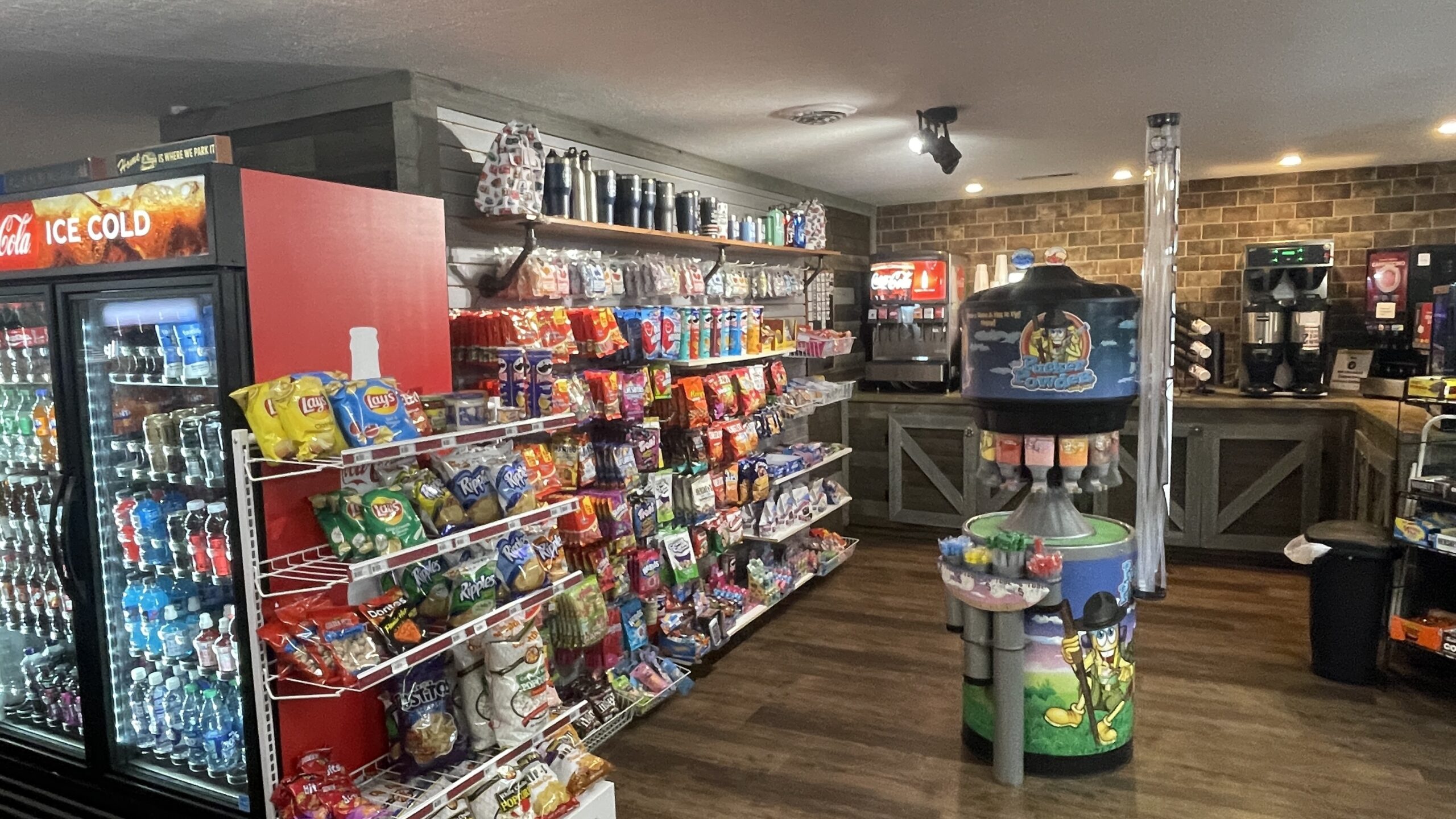 A convenience store interior with shelves of snacks and chips, coolers with drinks, coffee machines, and a slushie dispenser on a wood floor with brick and wooden walls.