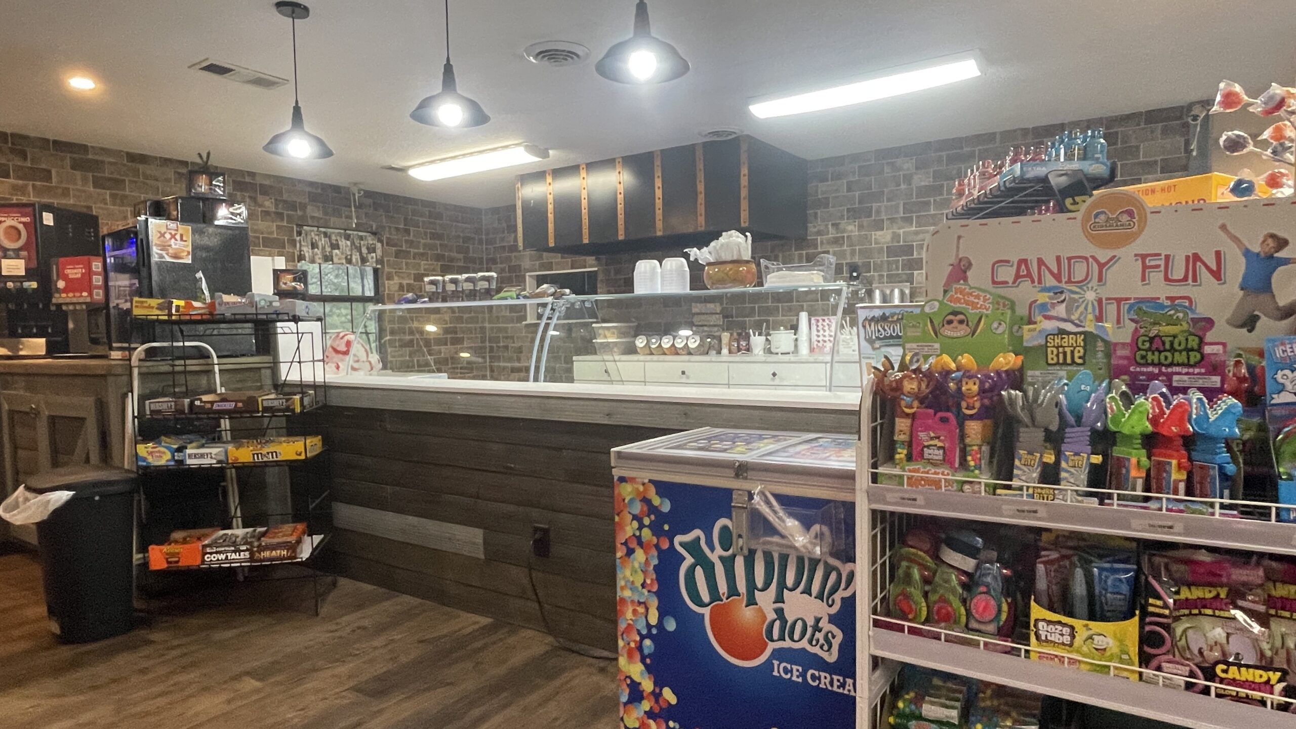 A brightly lit ice cream and candy shop interior features an ice cream counter, Dippin' Dots freezer, candy displays, snacks, and soda machines against a brick wall background.