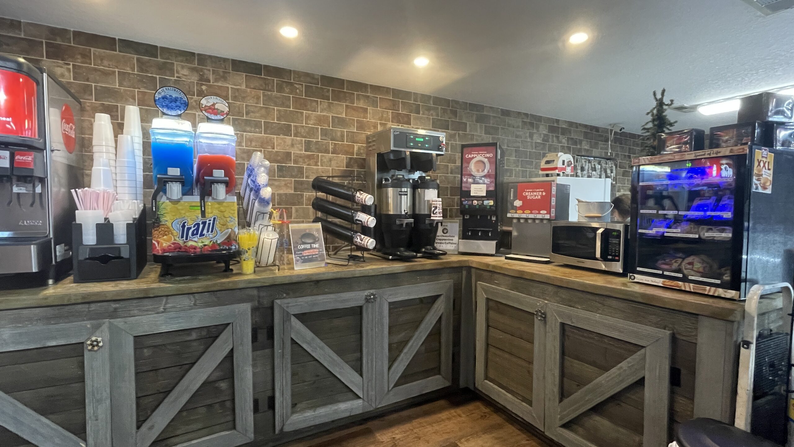A self-serve beverage station with soda dispensers, a juice machine, stacked cups, a coffee maker, a microwave, and a glass-front snack display, all against a brick wall with wooden cabinets below.
