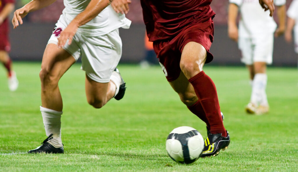 Two soccer players in motion, one wearing a maroon uniform and the other in white, compete for possession of a black-and-white soccer ball on a grassy field. Other players are visible blurred in the background.