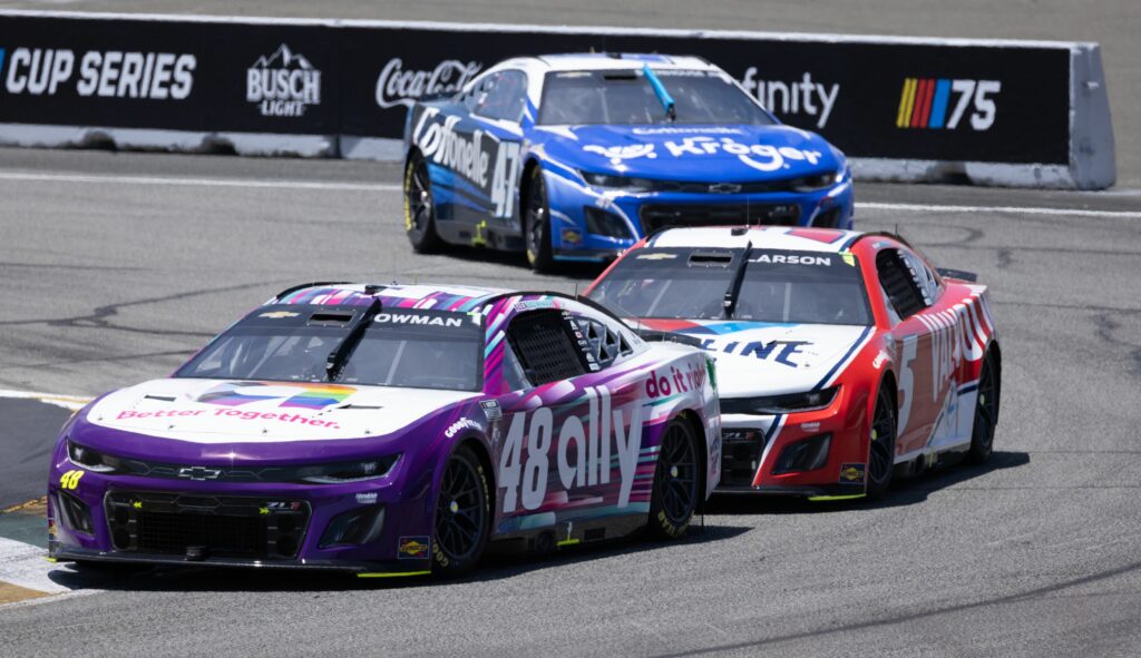 Three NASCAR race cars speed around a track. The front car is purple and white with the number 48, followed by a red and white car number 5, and a blue and white car number 47 in the background.