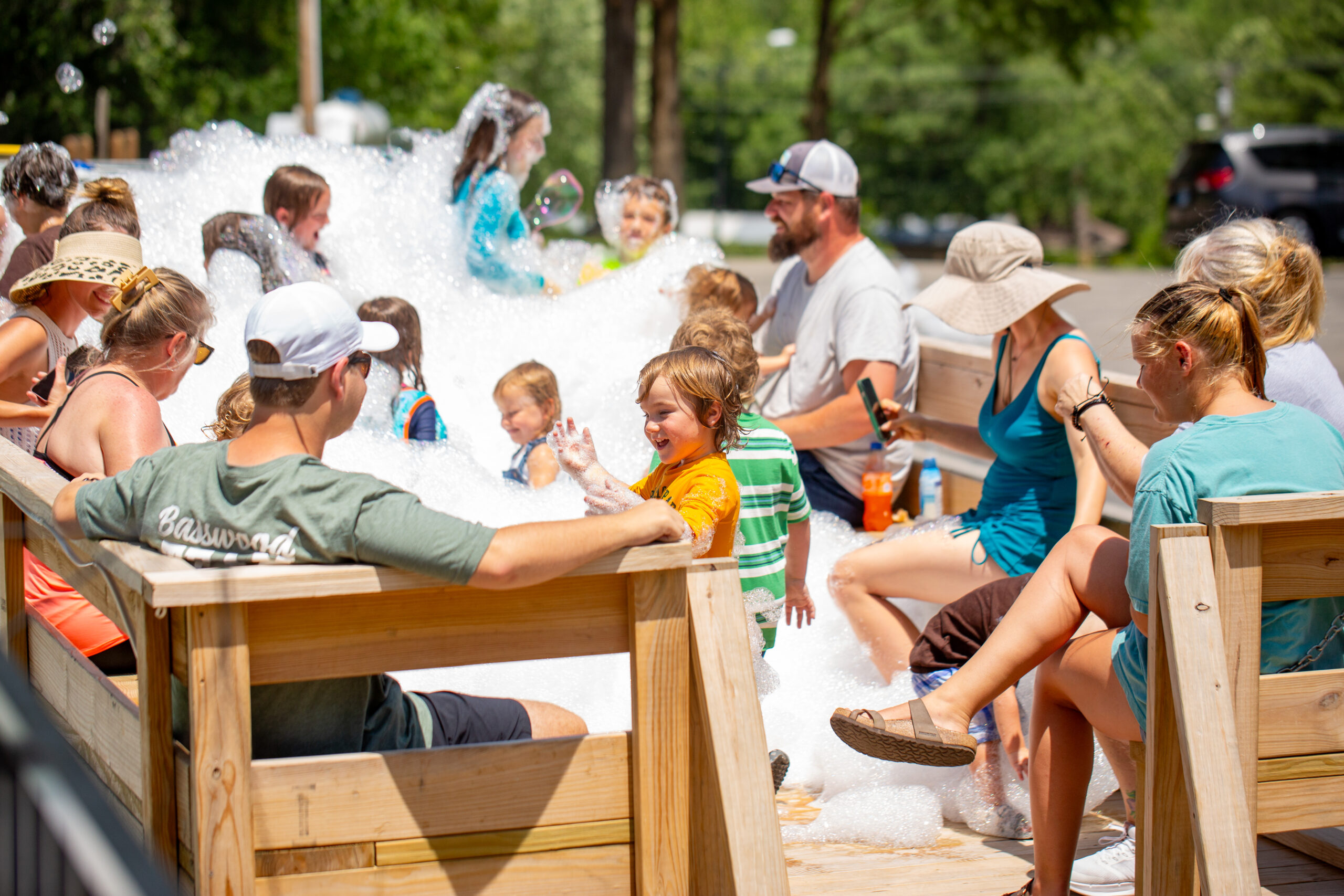 Children and adults enjoy a sunny day together on a wooden deck, with kids playing in a pile of bubbles while adults sit nearby, some wearing hats and sunglasses. Trees and a parking lot are visible in the background.