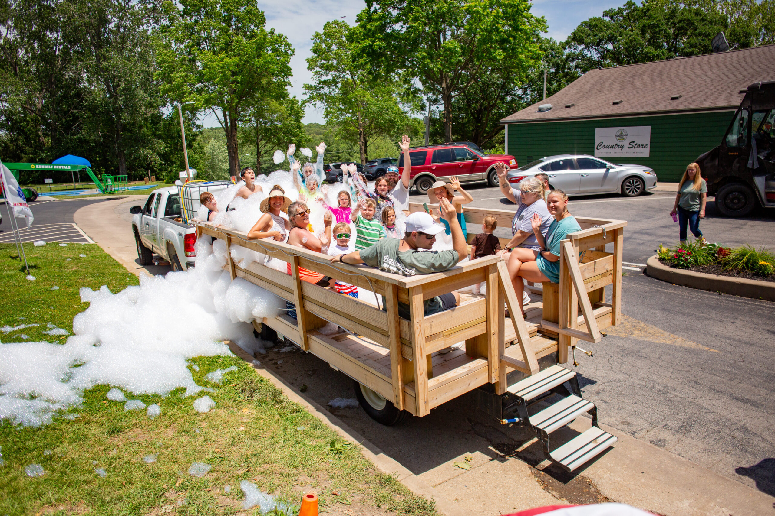 A group of people, including children and adults, ride on a wooden trailer covered in soap bubbles, being pulled by a white truck near a green building on a sunny day. Most are smiling and waving.