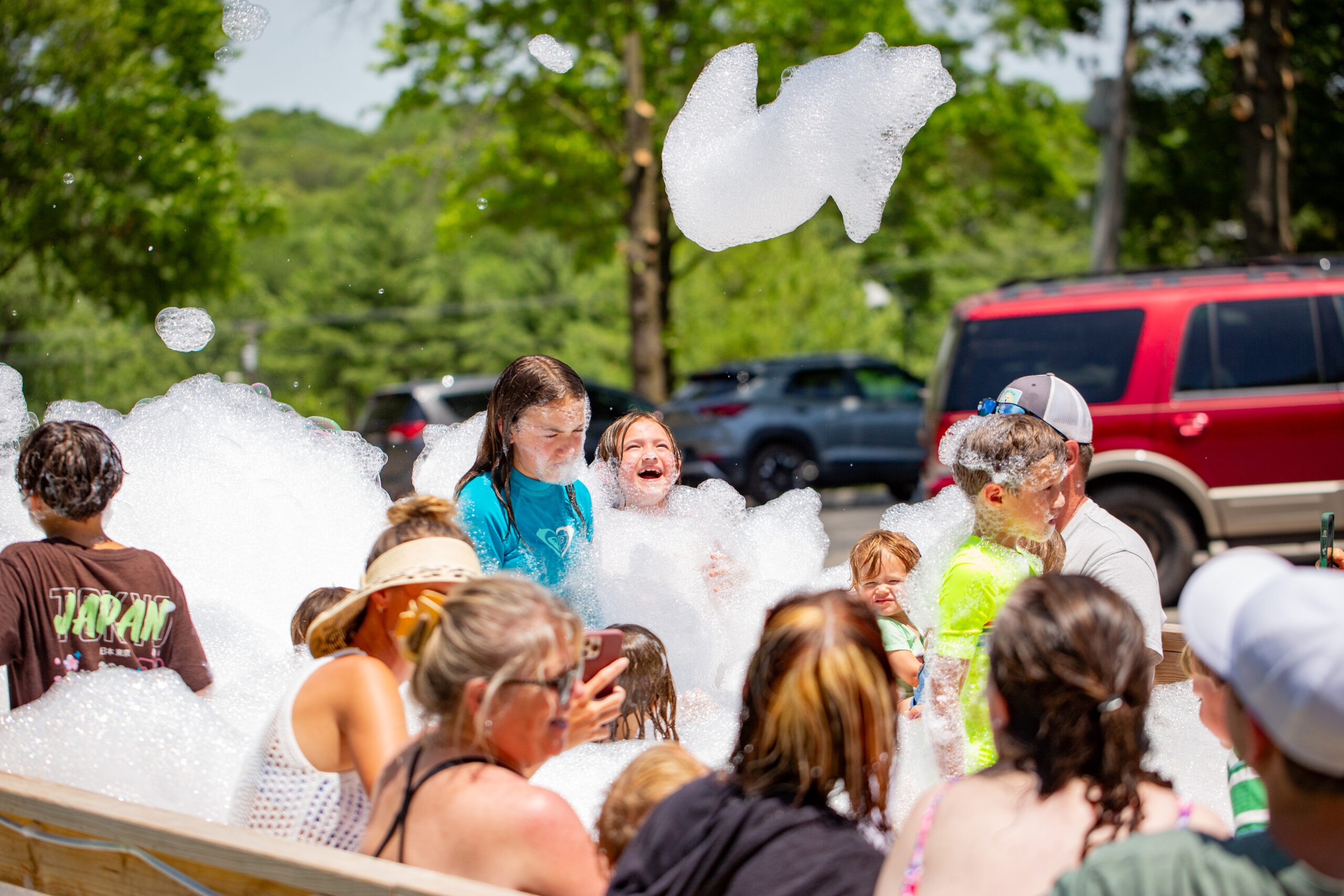 A group of children and adults play outside in thick white foam on a sunny day, laughing and smiling. Trees and parked cars are visible in the background. A large clump of foam floats in the air above them.