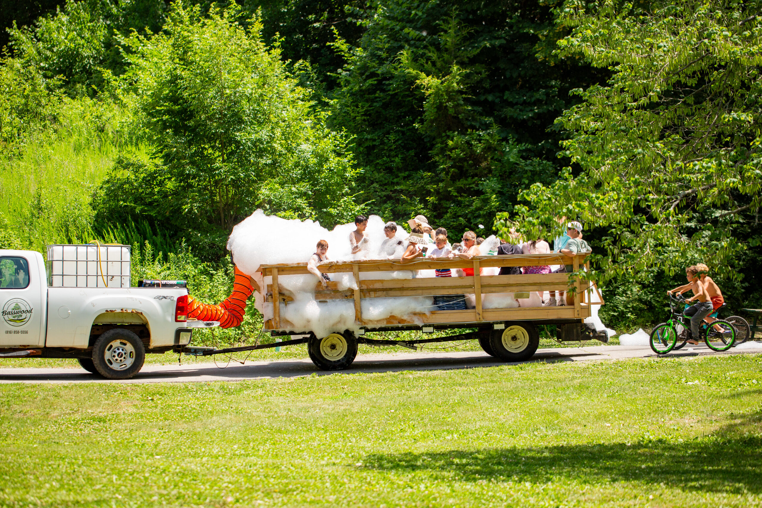 A pickup truck pulls a wooden trailer filled with children playing in foam. The trailer is surrounded by greenery, and two children on bikes ride nearby on a sunny day.