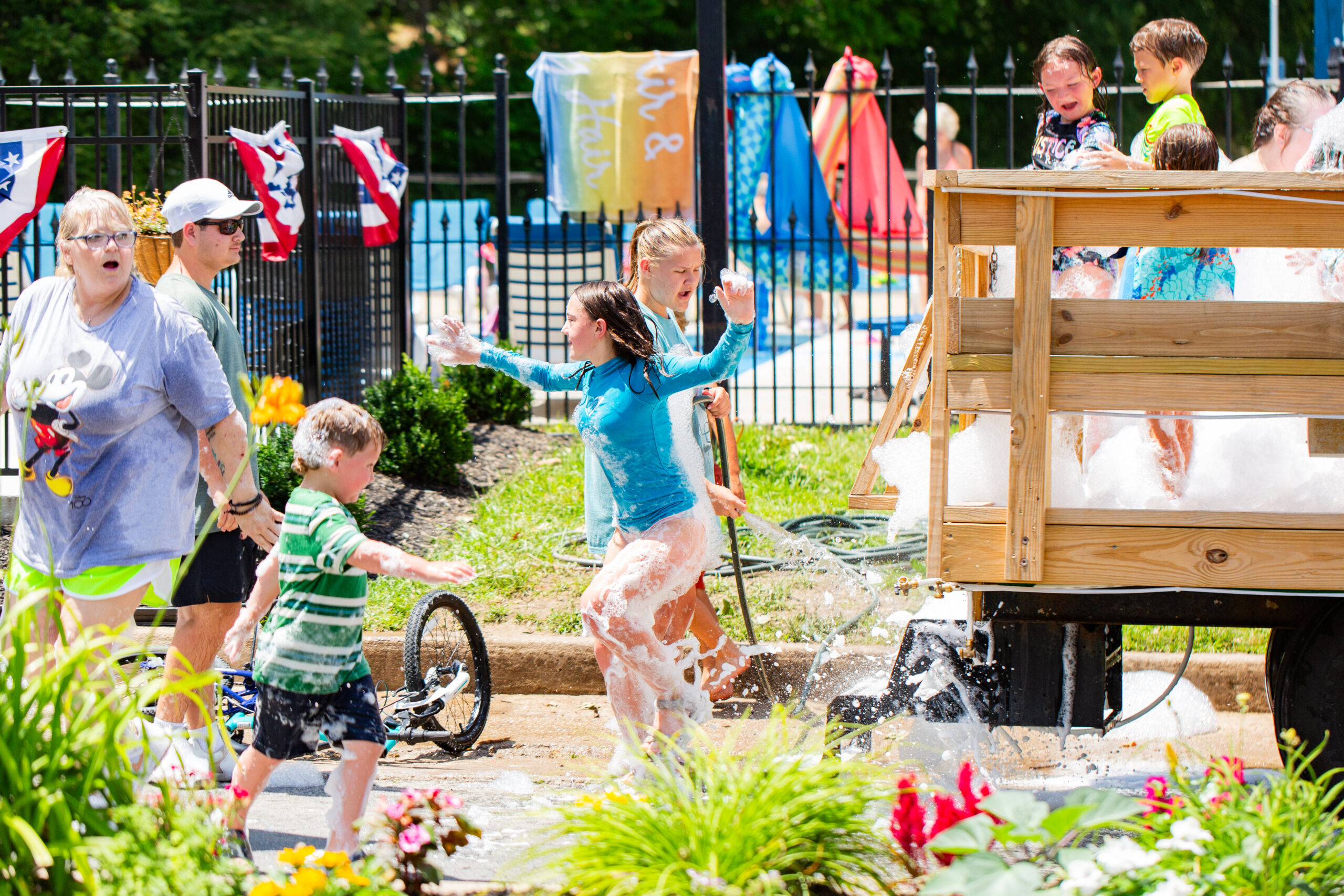 Children and adults play and run around a wooden cart filled with soapy water, splashing and laughing outdoors near a pool on a sunny day. Bright towels and decorations are visible in the background.