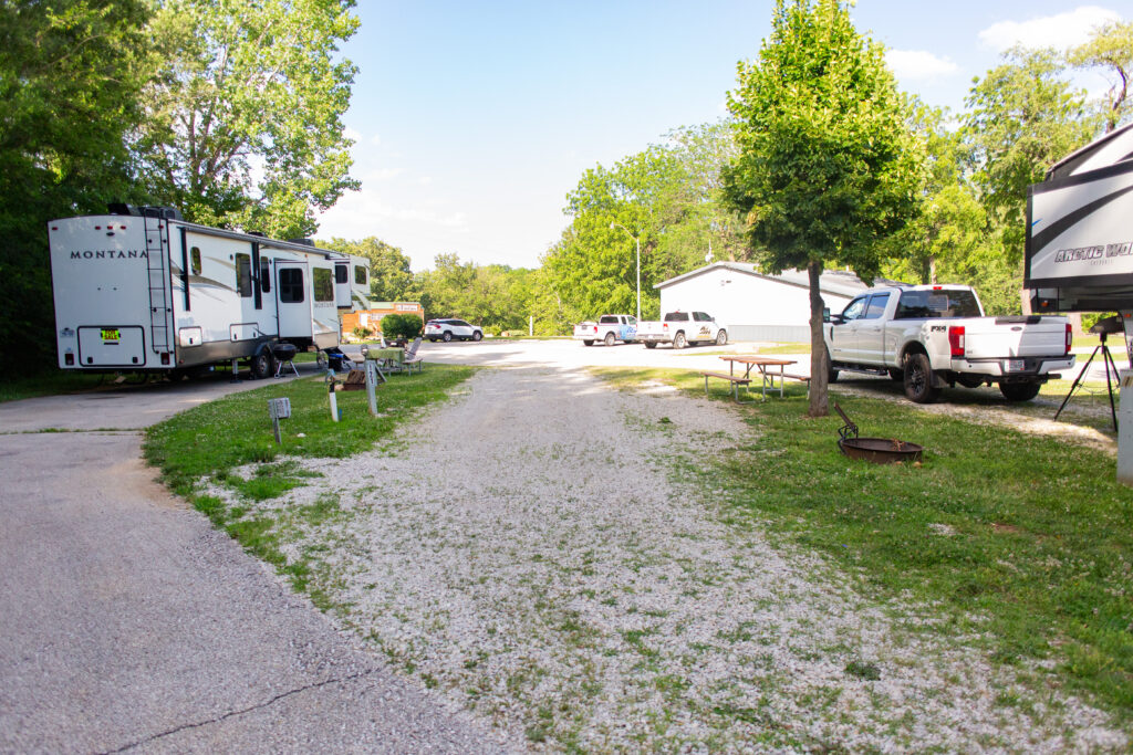 A gravel road runs through a campground with RVs, trucks, picnic tables, and a fire ring surrounded by grass and trees under a partly cloudy sky.