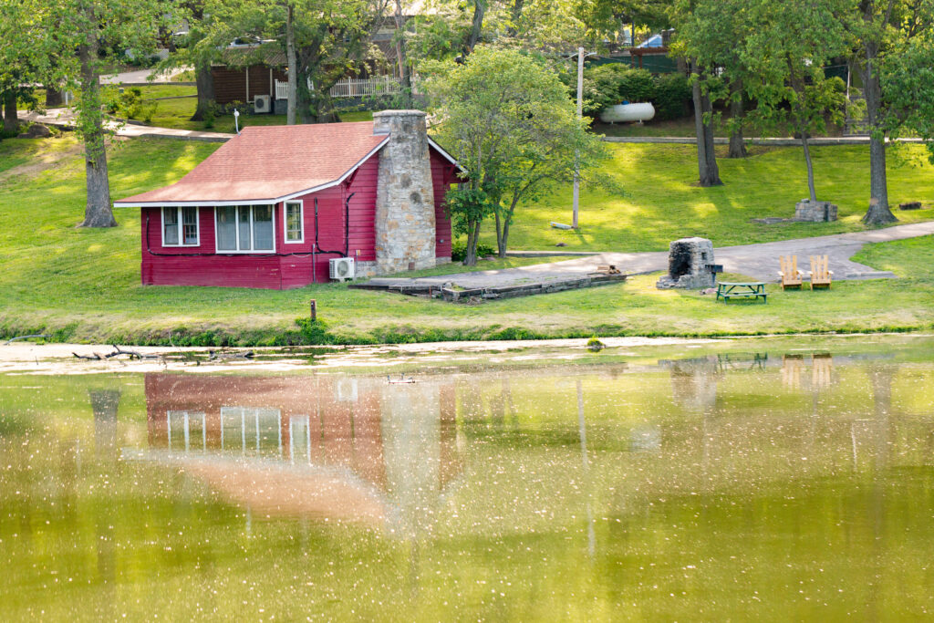 A small red cabin with a stone chimney sits on a grassy hill by a calm lake, with its reflection visible in the water. Trees and outdoor seating surround the cabin on a sunny day.