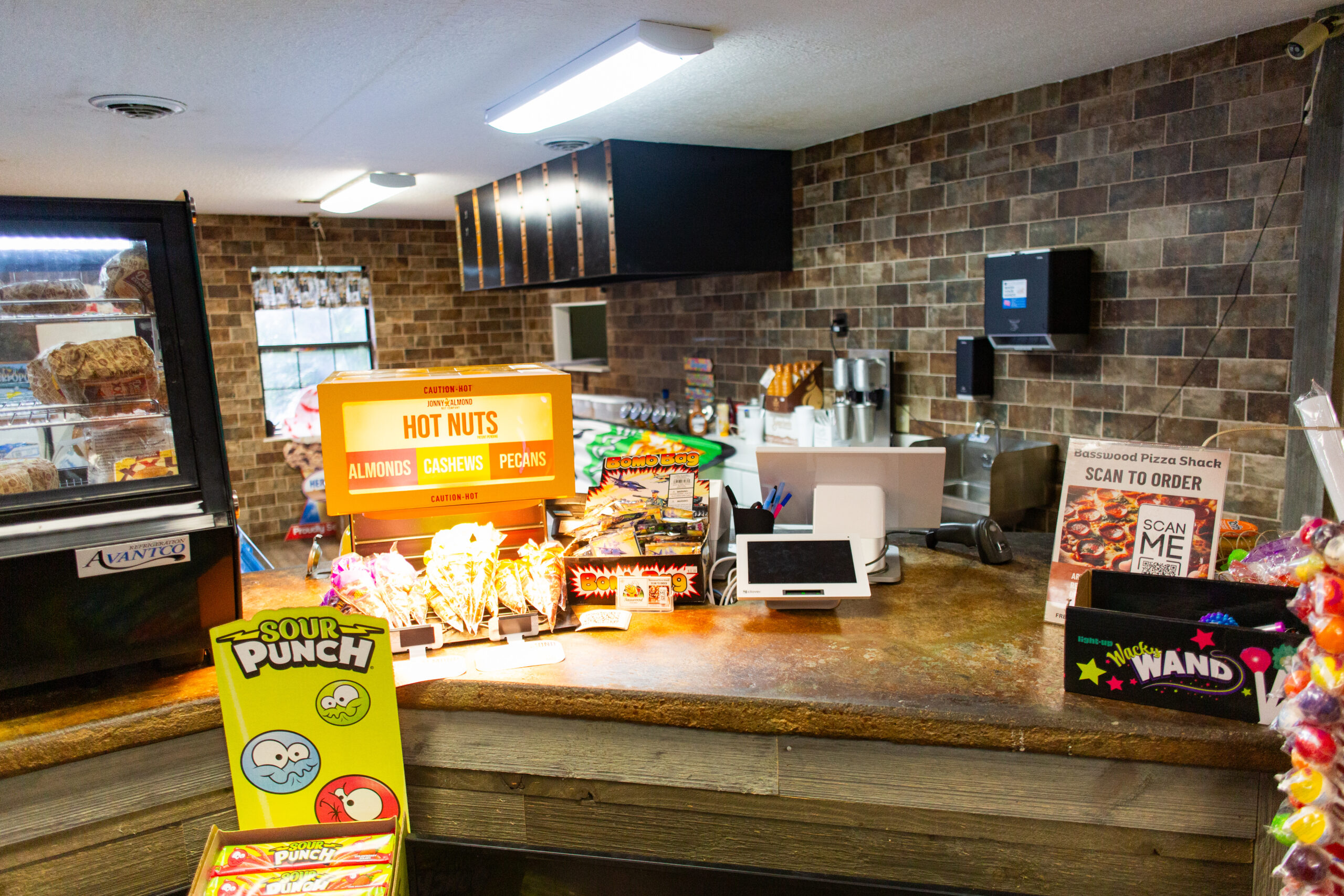 A counter in a snack shop displays hot nuts, candy, a cash register, a QR code sign, and a glass case with baked goods, with brick walls and bright lighting.
