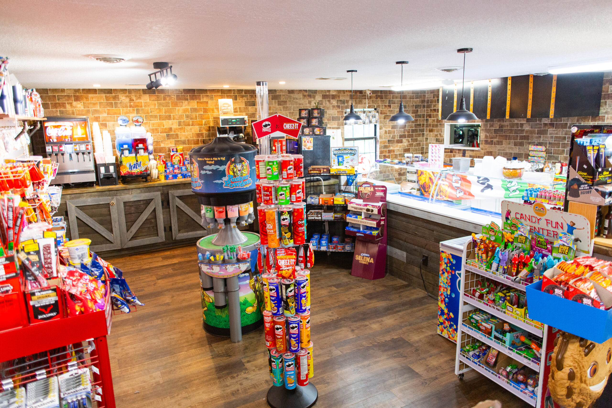 A bright, colorful candy store interior with shelves and racks stocked with candy, snacks, and drinks. The wooden floor and brick walls add a cozy feel. Various treats are neatly displayed throughout the store.