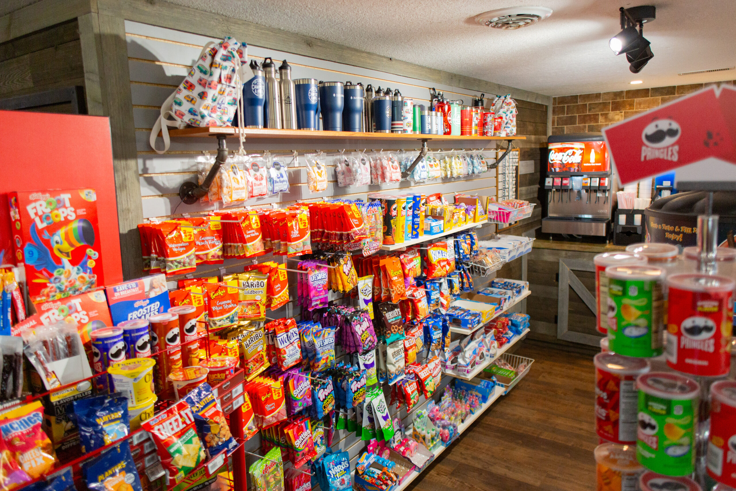 A brightly lit snack shop with shelves filled with chips, candy, and snacks; travel mugs and water bottles are displayed above, and a soft drink machine is visible in the background.