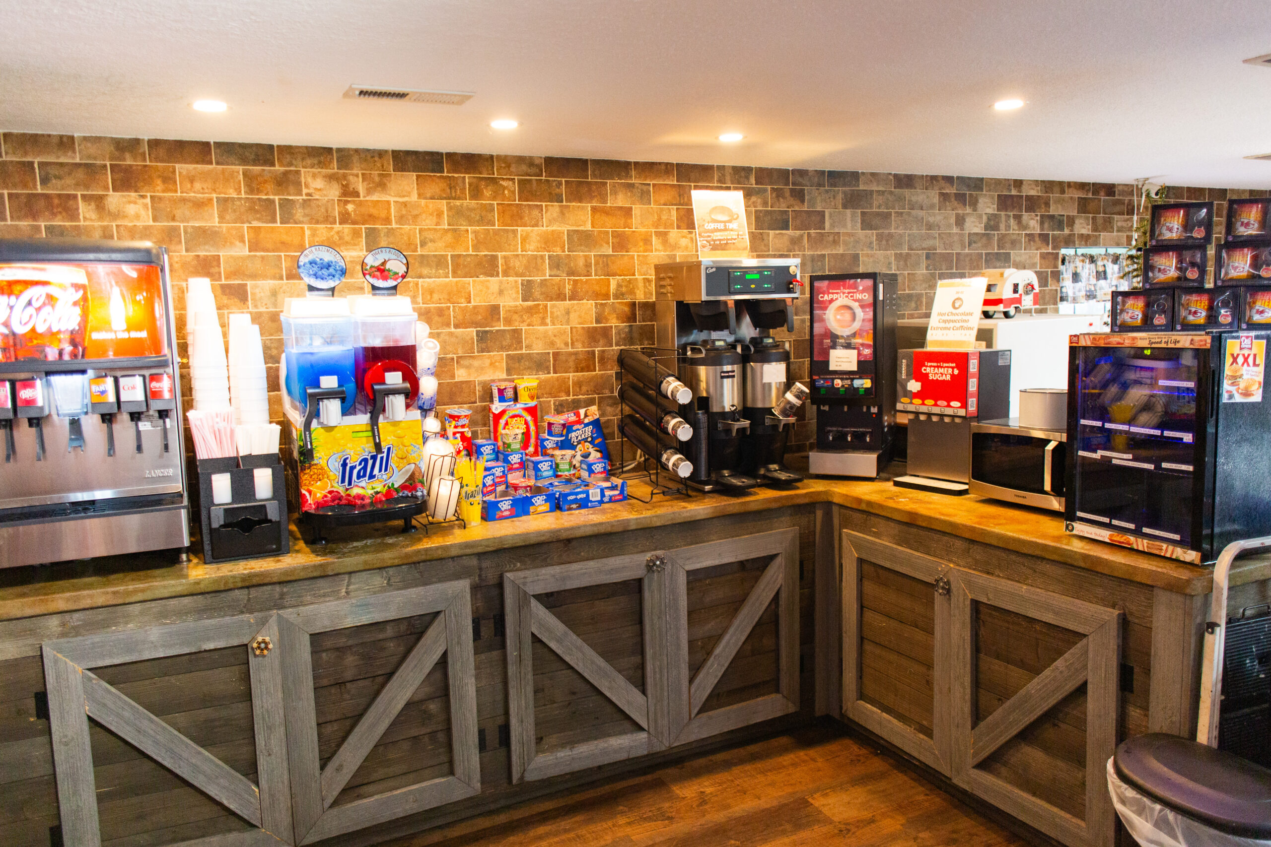 A self-serve beverage and snack station with soda fountains, slushie machines, coffee dispensers, cups, and assorted snacks on a wooden counter against a tiled wall.