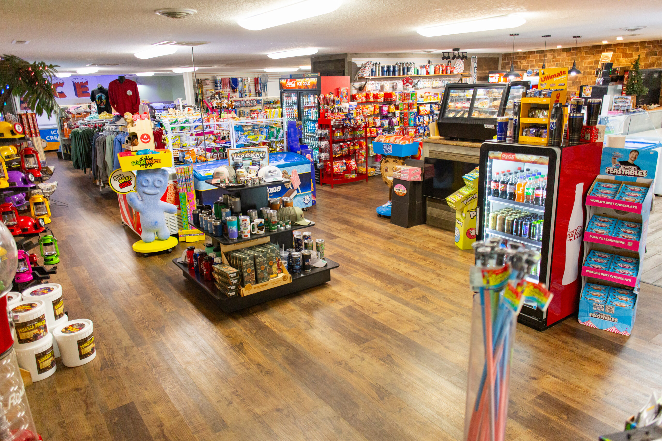 Brightly lit convenience store with wooden floors, colorful snacks, drinks, household items, and toys on shelves. A blue cartoon character statue stands near the center, and freezers and refrigerators line the walls.