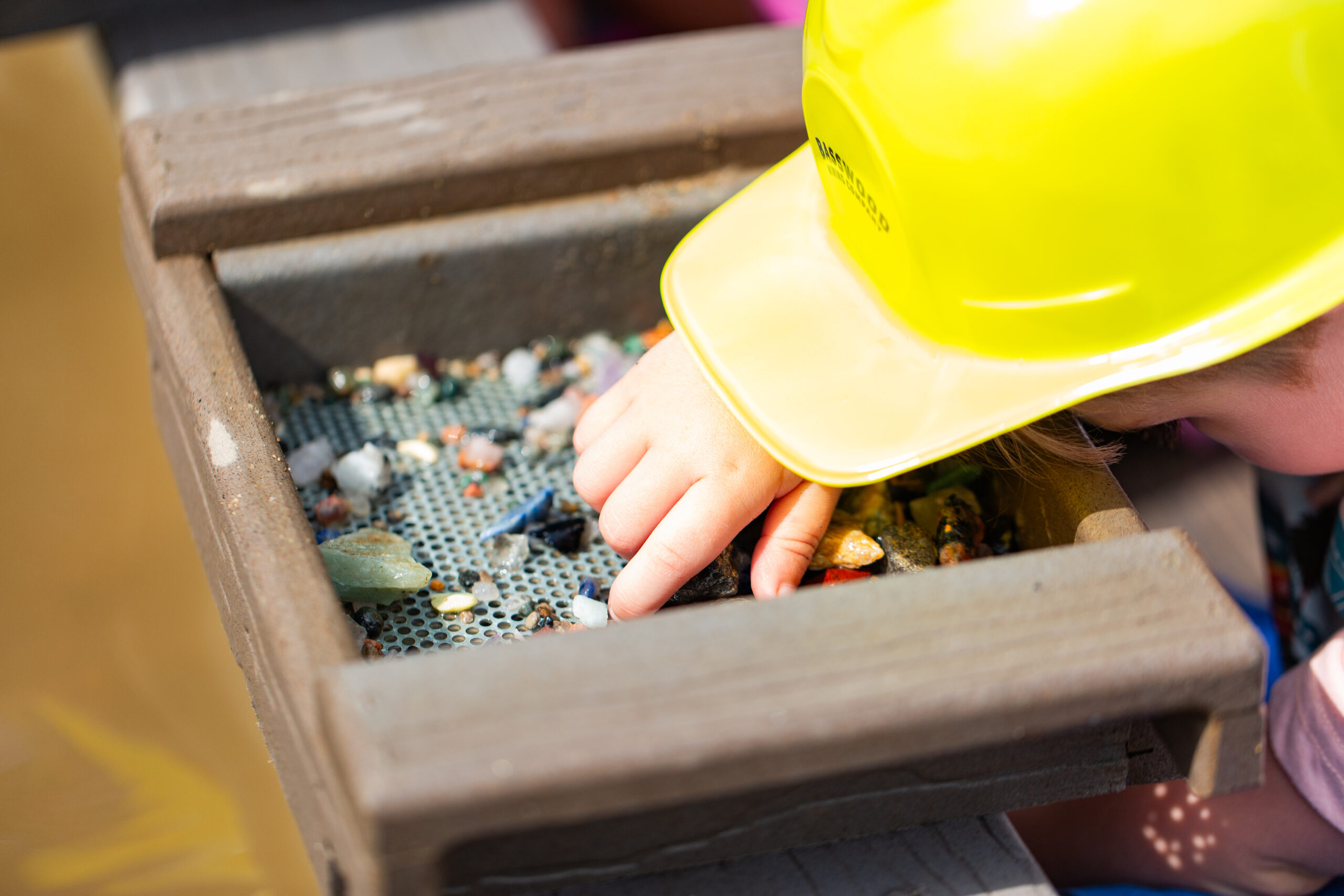 A child wearing a yellow construction helmet sifts through small rocks and gems in a wooden tray, using their hand to search through the materials.