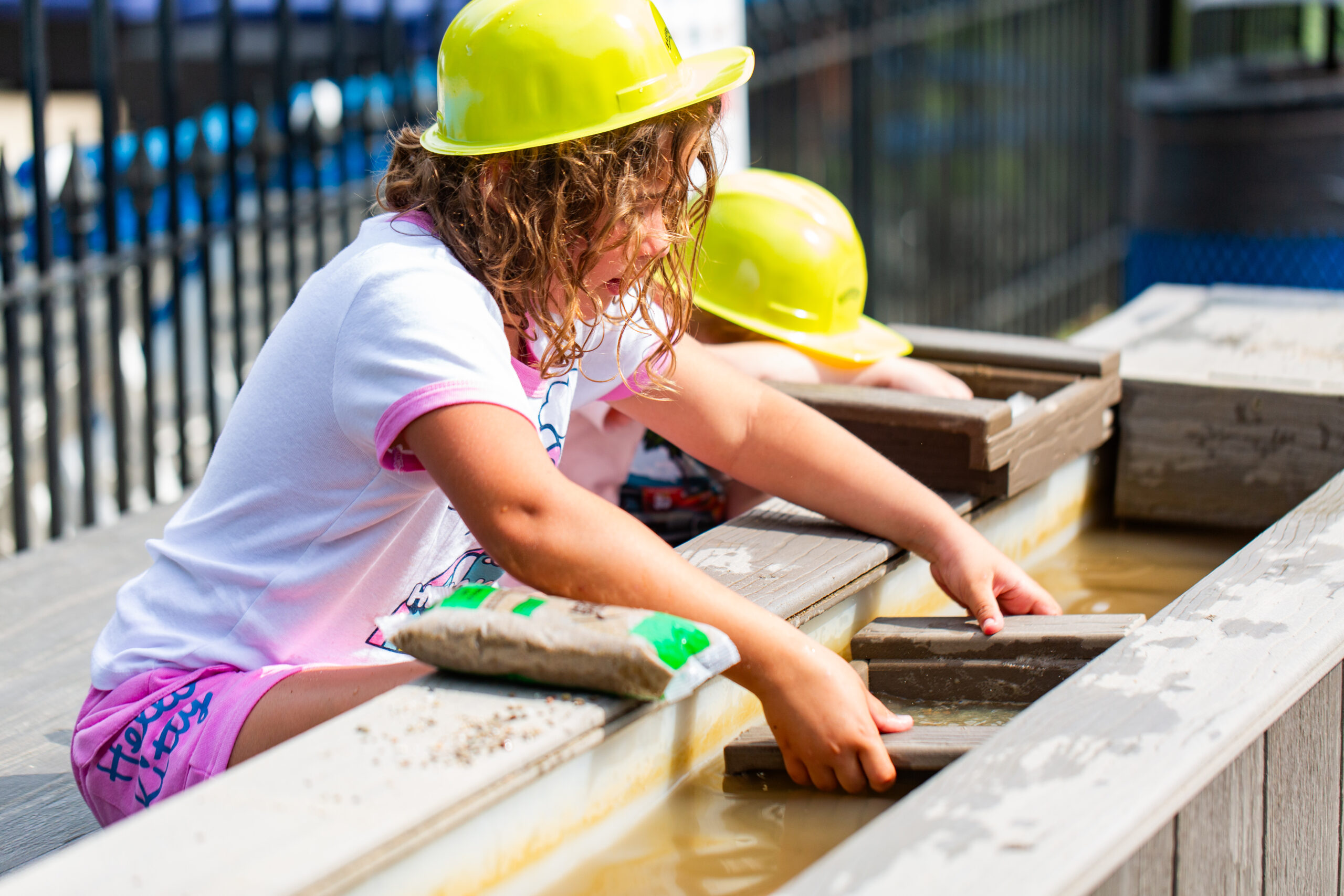 A young child wearing a yellow hard hat plays with water and sand at an outdoor activity station, using wooden trays to sift through muddy water. Another child in a hard hat is partially visible beside her.