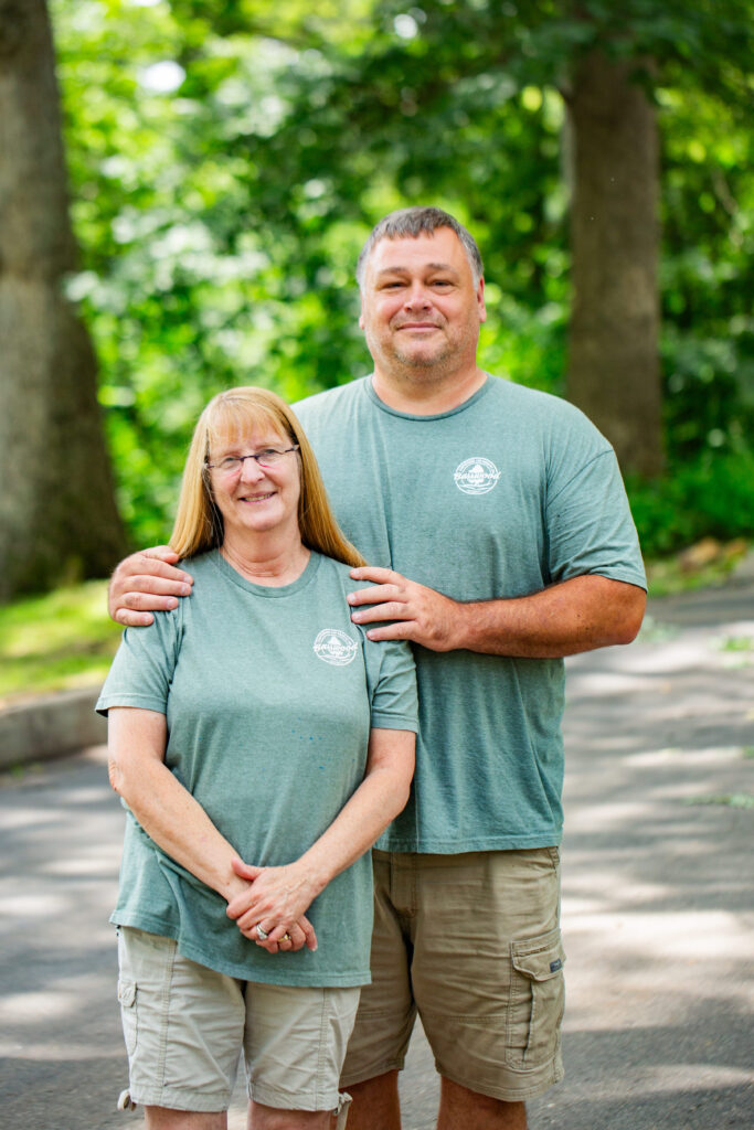 A woman and a man stand outdoors on a sunny day, both wearing matching green T-shirts and khaki shorts. The man stands behind the woman with his hand resting on her shoulder. Trees and greenery are visible in the background.