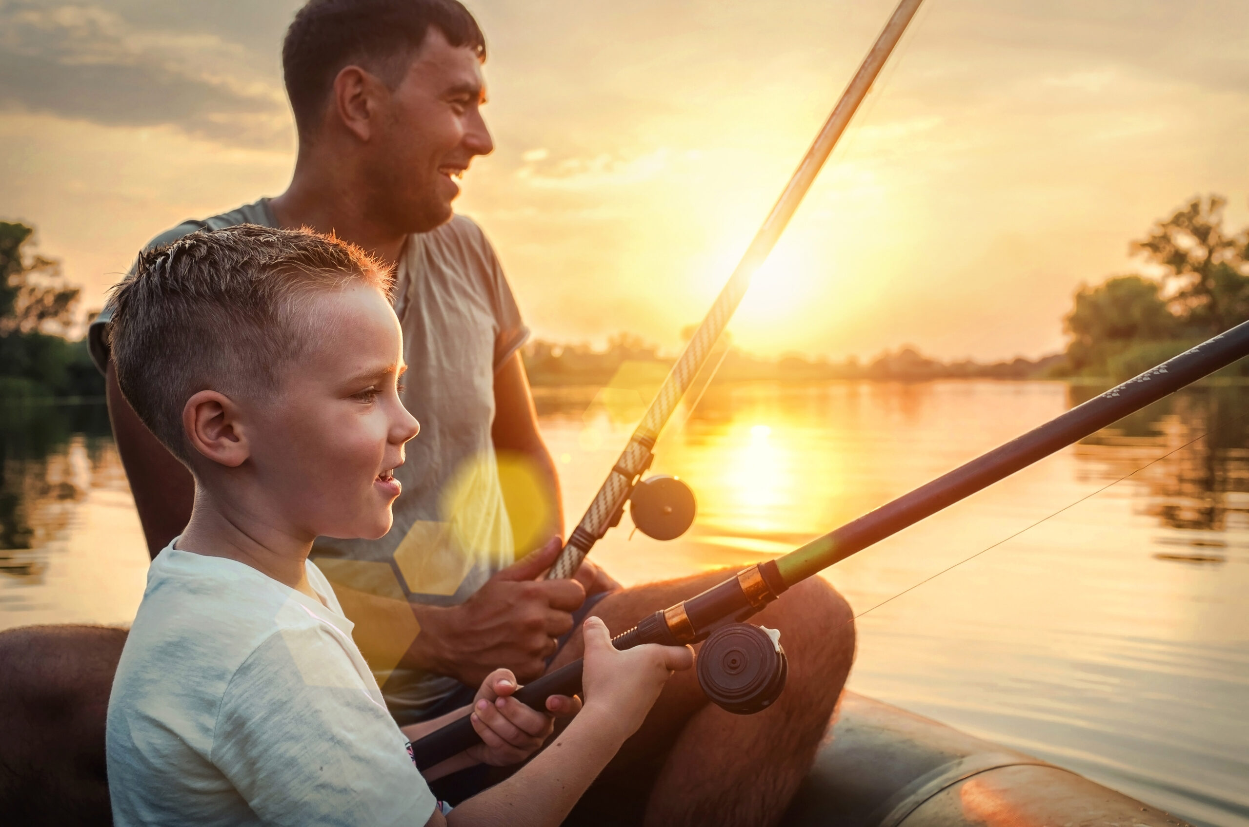 A young boy and an adult, both smiling, are fishing together on a boat at sunset. The sun is low over a calm lake, casting a warm glow on the water and their faces. Trees line the distant shore.