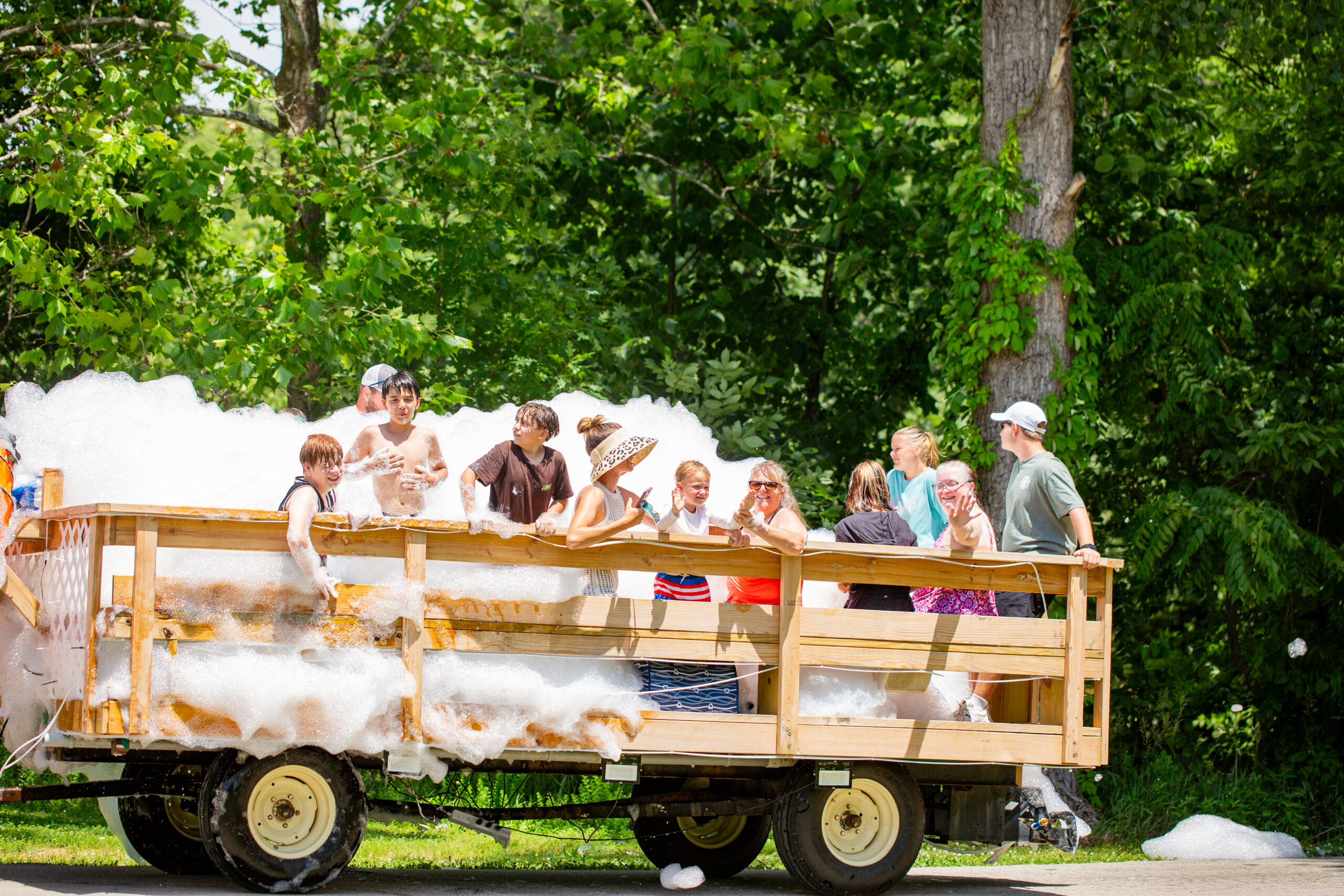 A group of children and adults stand on a wooden trailer filled with foam, smiling and having fun in a lush, green outdoor setting with trees in the background—perfect for any summer event photo gallery.