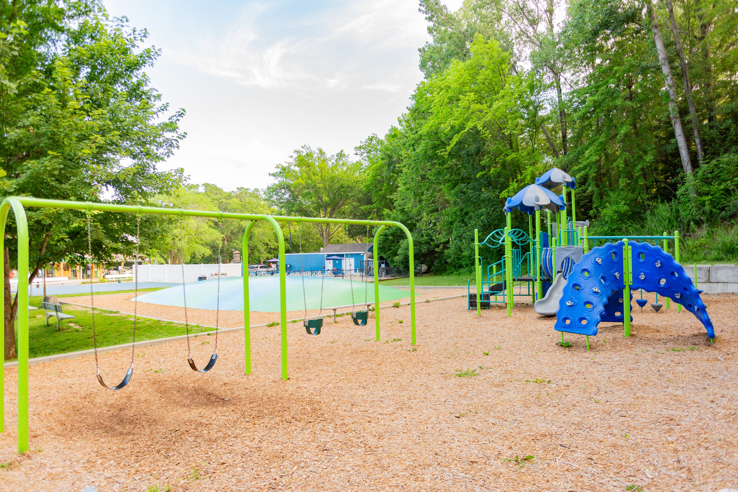 A Photo Gallery-worthy playground with green swings, a blue climbing structure, and a slide sits among trees and wood chip ground cover on a sunny day. A splash pad and park buildings are visible in the background.
