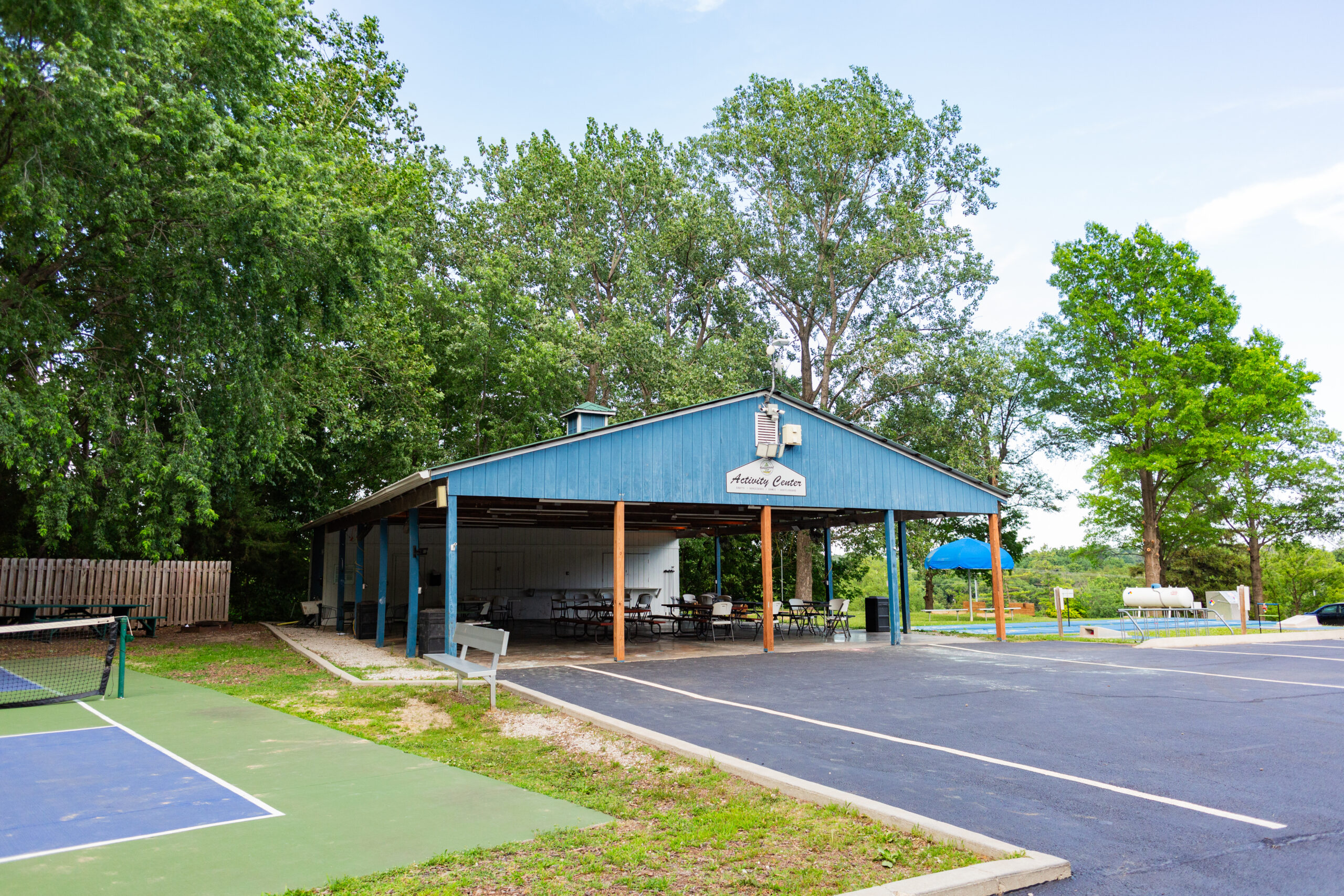 A covered picnic pavilion with tables sits next to a parking lot, surrounded by trees. A tennis court is visible in the foreground, while a blue playground structure appears in the background. Explore more views like this in our Photo Gallery.