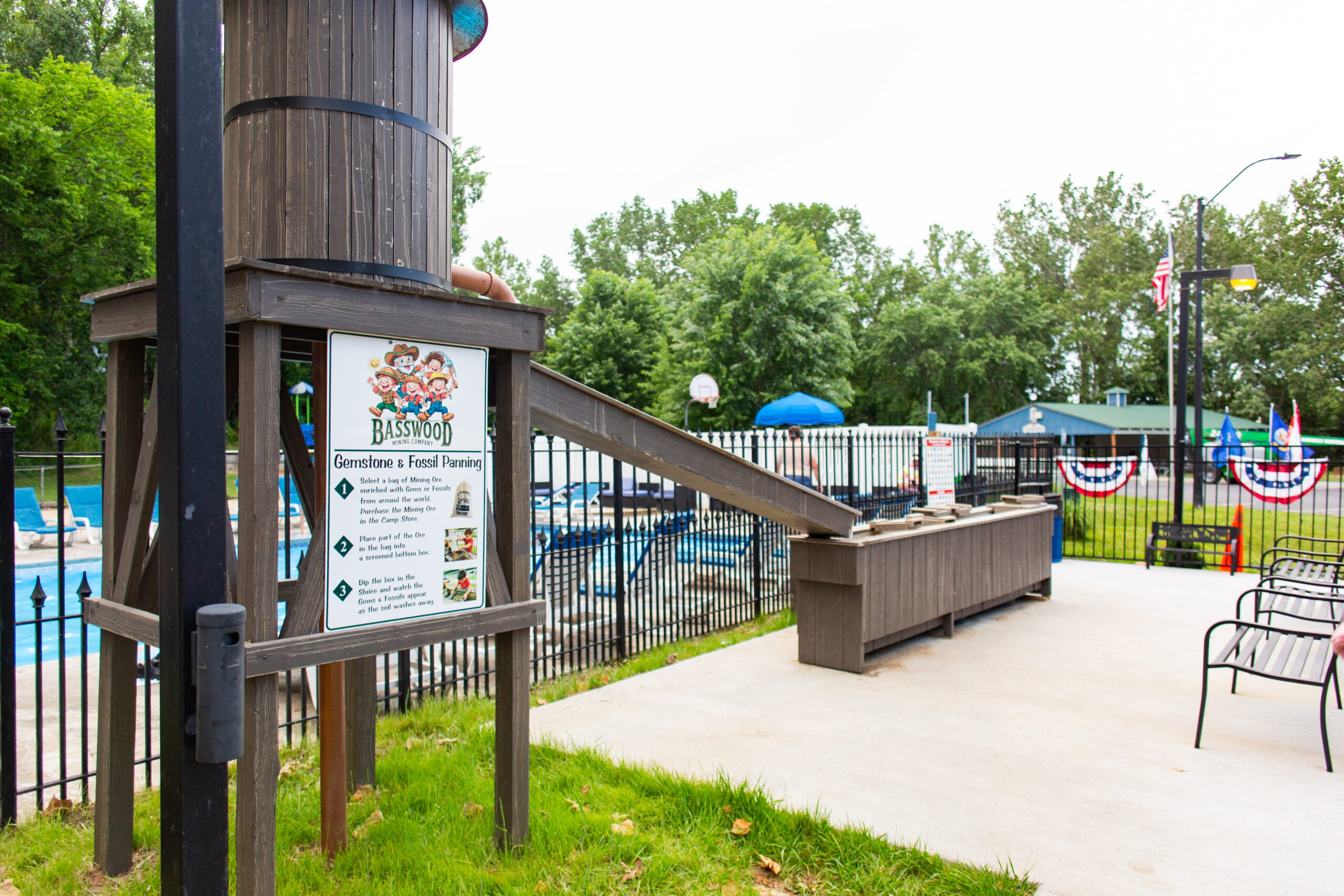 Outdoor gem mining station near a fenced pool, with a wooden water tower, display sign, sluice troughs, and benches. Trees and picnic tables are visible in the background. American flag bunting lines the fence—see more in our Photo Gallery.