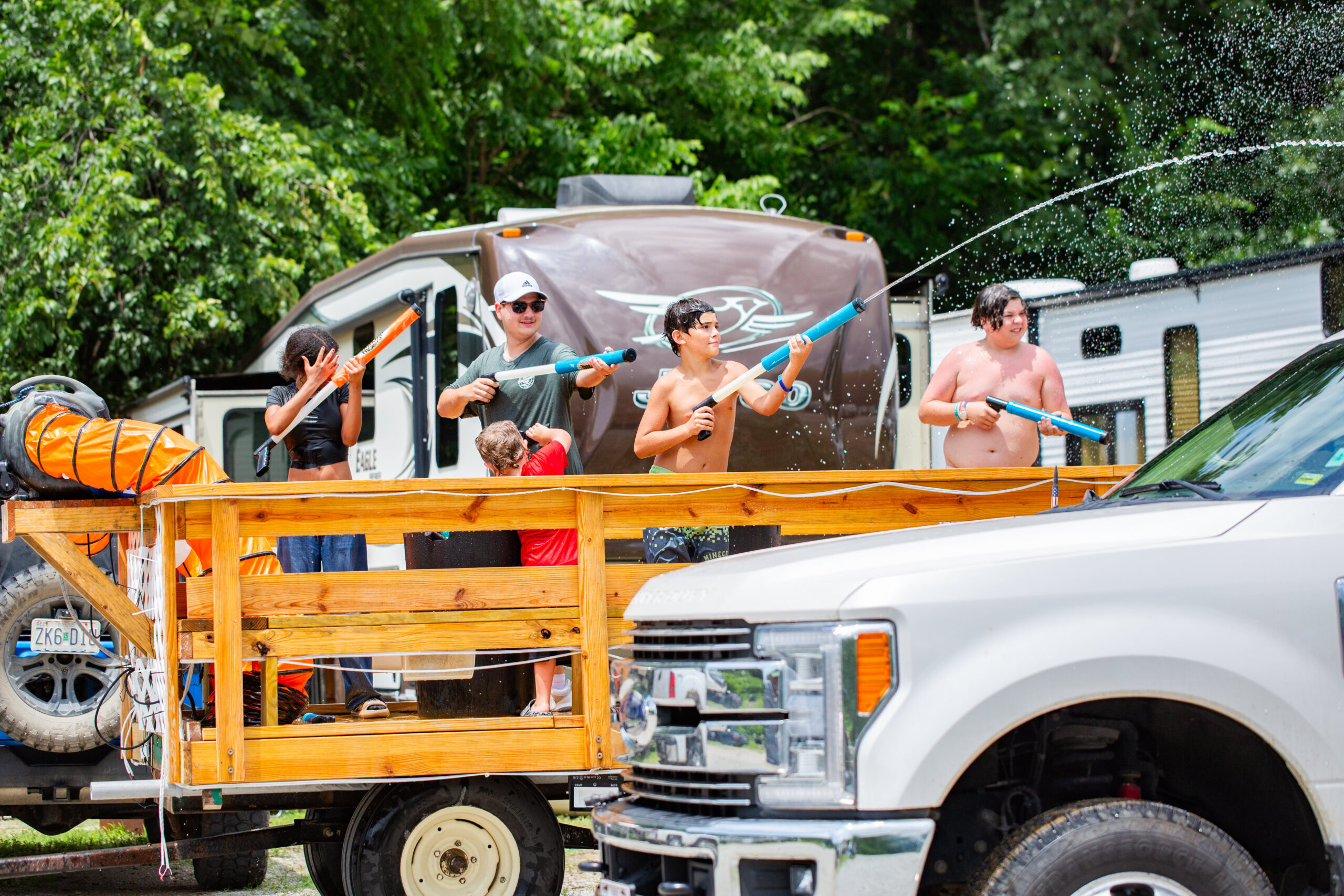 In this lively photo gallery moment, five kids, some shirtless, stand on a wooden trailer with water guns, spraying a white pickup truck. An RV and trees set the sunny-day backdrop.