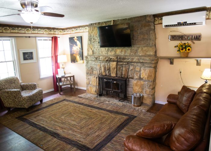 Cozy living room with stone fireplace, mounted TV, leather sofa, armchair, large rug, windows with red and white curtains, side tables with lamps, and a “Welcome to the Lake” sign on the wall.