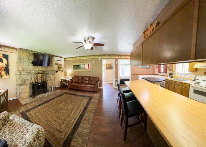 Wide-angle view of a cozy living room with brown sofas, a stone fireplace, wall-mounted TV, and a breakfast bar overlooking a kitchen with white appliances and wooden cabinets. Warm lighting and homey décor throughout.