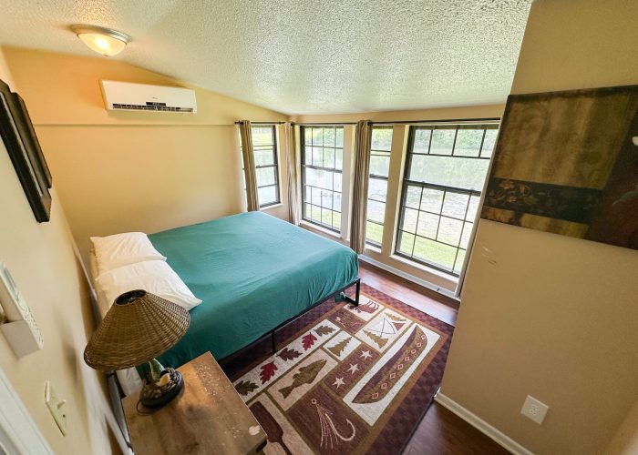 A cozy bedroom with a green bedspread, a wooden nightstand with a lamp, large windows letting in natural light, and a patterned rug on the floor. The room features beige walls and dark wood flooring.
