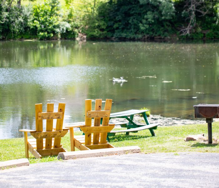Two wooden chairs face a calm pond, perfect for a long-term stay, with a green picnic table and metal grill nearby. Trees and greenery line the opposite shore under bright sunlight.