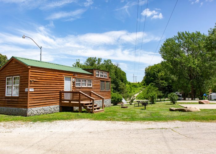 A small wooden cabin with a green metal roof sits on a grassy lot with trees and picnic tables nearby under a blue sky with wispy clouds. A gravel road runs in front of the cabin.