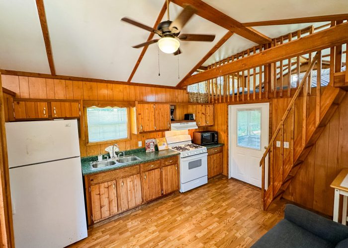 A cozy wooden cabin kitchen with a white fridge, sink, stove, and microwave. Stairs lead to a loft above, and there is a ceiling fan and a door with a window. The space has wood-paneled walls and floors.