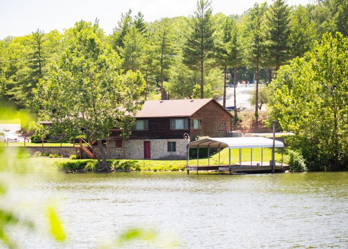 A wooden cabin with a stone foundation sits on the shore beside a lake, surrounded by green trees. A covered dock extends into the water in front of the cabin under a sunny sky.