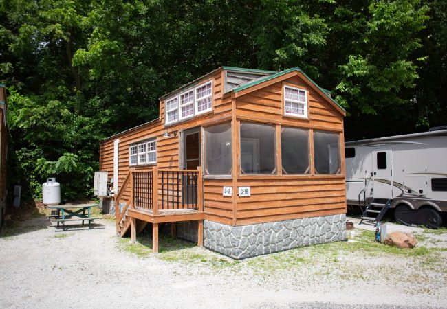 A small wooden cabin with a screened porch, stone foundation, and nearby picnic table, situated next to a white travel trailer and surrounded by trees.