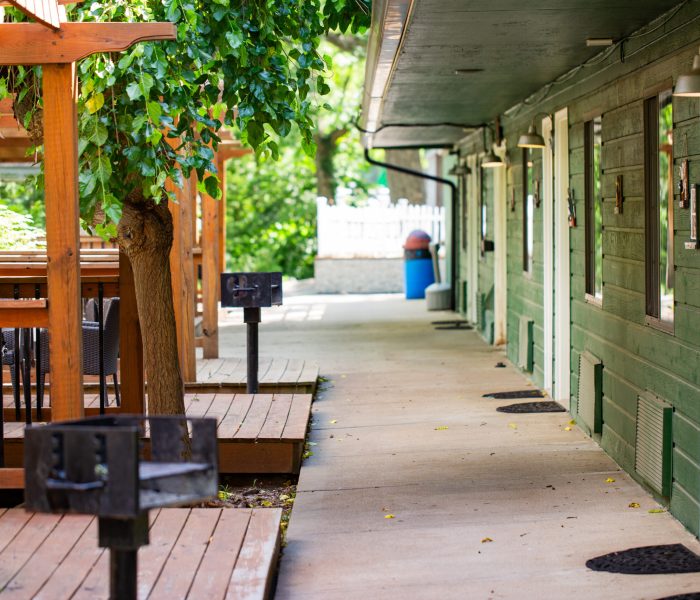 A shaded outdoor walkway beside a green wooden building offers picnic tables and grills on wooden decks—perfect for a relaxing long-term stay, with leafy trees overhead and a white picket fence in the background.