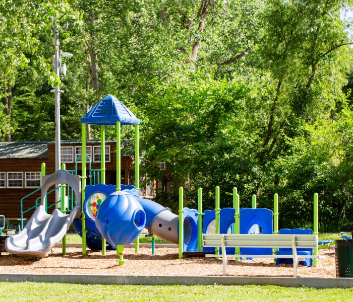 A colorful playground with blue and green slides, tunnels, and climbing structures set on wood chips is surrounded by trees and greenery—perfect for long-term stay visitors—with a bench and a building in the background.