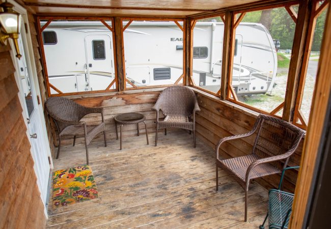 A small screened porch with wicker chairs, a table, and a colorful doormat overlooks a parked travel trailer.