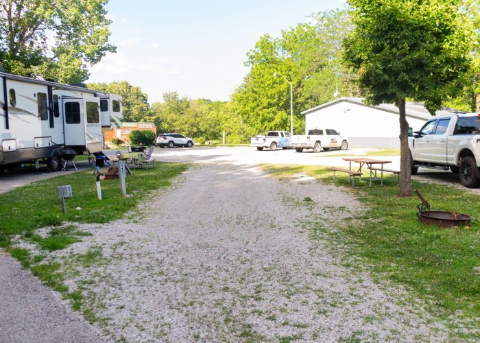 A gravel campsite with picnic table and fire ring, surrounded by green grass and trees; RVs and parked cars are visible on either side under a clear blue sky.
