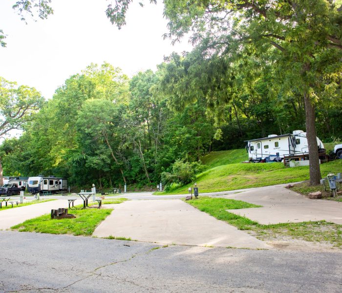 A campground with empty concrete RV pads, picnic tables, and hookups, surrounded by green trees. Several RVs are parked along the side, and the area is bright with natural sunlight.