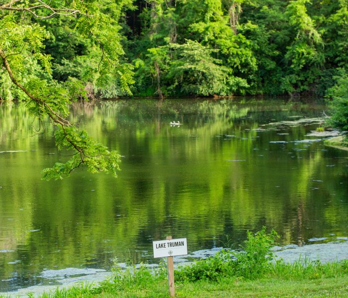 A serene pond surrounded by lush green trees, perfect for a long-term stay, with a small sign in the grass reading "Lake Truman" near the water's edge. The calm lake reflects the trees beautifully.