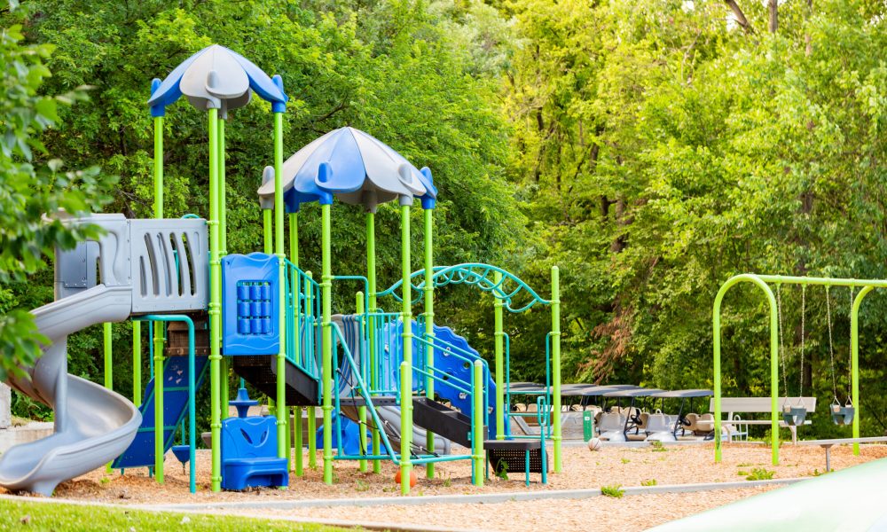 A colorful playground with blue and green climbing structures and slides stands empty, surrounded by trees and greenery. Swings, benches, and picnic tables are also visible in the background.