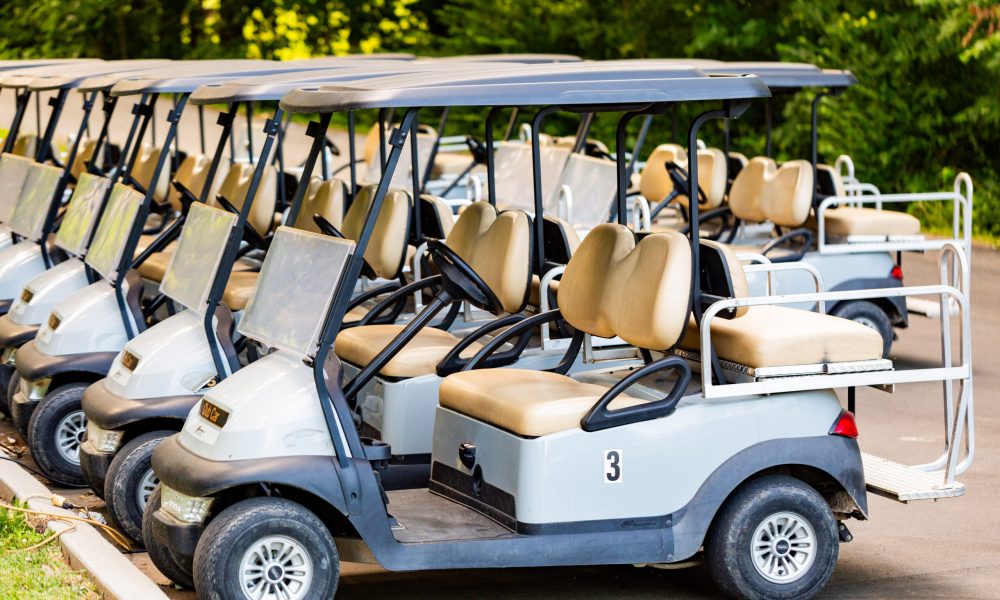A row of parked golf carts with beige seats and white exteriors lined up on a paved area, surrounded by green trees in the background.