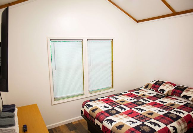 A small, simple bedroom with white walls, a bed with a wildlife-themed quilt, a TV on the wall, a window with closed blinds, and a wooden desk with folded towels and a remote.