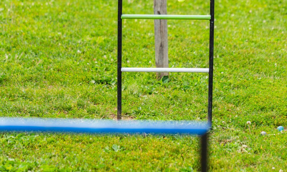 A ladder golf game setup on green grass, with bolas hanging on the middle rung and a scoreboard in the background. The focus is on the ladder, with another ladder blurred in the foreground.