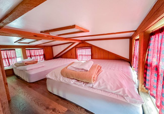 Two twin beds with white bedding and folded towels are placed in a small loft room with wooden beams, wood walls, and red plaid curtains covering the windows.